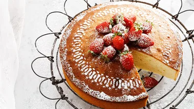 Strawberry-topped sponge cake with powdered sugar on a decorative wire stand, one slice missing.