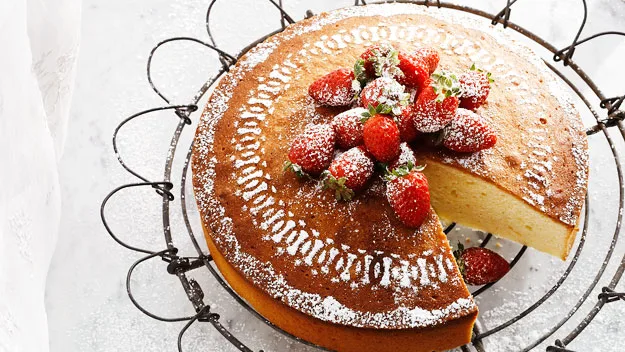 Strawberry-topped sponge cake with powdered sugar on a decorative wire stand, one slice missing.