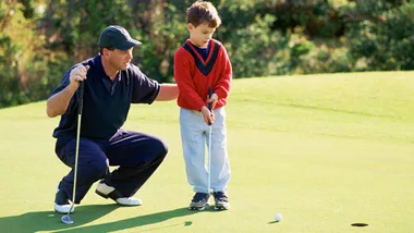 Man guides young boy in red sweater on golf putting green, teaching him how to putt a golf ball into the hole.