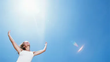 "Woman in a white dress joyfully reaching towards the bright sun in a clear blue sky."