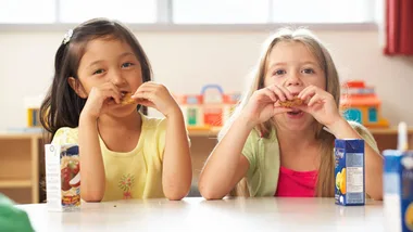 Two young girls sitting and eating snacks at a table with juice boxes beside them.