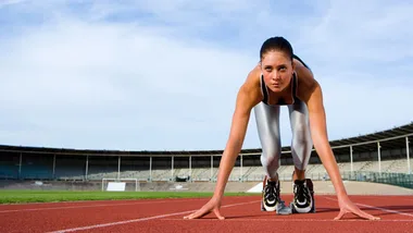 Female athlete in start position on a track, wearing black shoes and white leggings, with a stadium in the background.