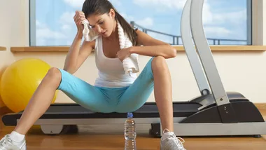 Woman resting on a treadmill after a workout, wiping sweat with a towel, with a water bottle and exercise ball nearby.