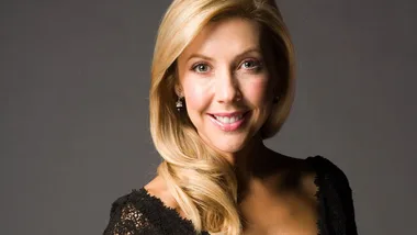 A woman with long blonde hair and a black lace dress smiles at the camera against a gray background.