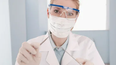 Dentist wearing safety goggles, mask, and gloves, holding dental tools, standing in a light blue dental office.