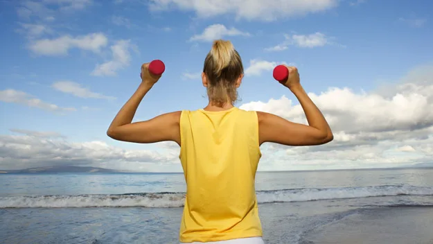 Woman exercising with dumbbells on a beach, facing the ocean under a blue sky.