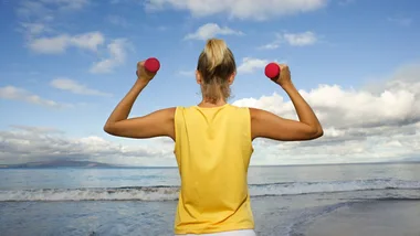 Woman exercising with dumbbells on a beach, facing the ocean under a blue sky.