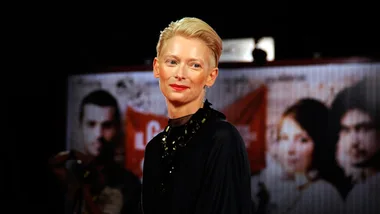 Tilda Swinton smiling at an event, with a poster featuring actors in the background.