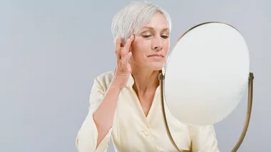 Mature woman with short gray hair examining her skin in a handheld mirror against a neutral background.