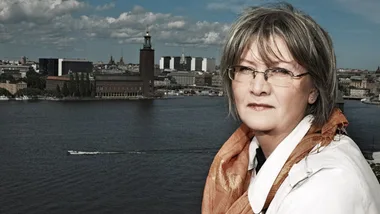 A person with glasses and scarf stands outdoors with a city skyline and waterway in the background.