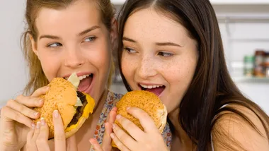 Two women smiling and holding cheeseburgers, about to take a bite.