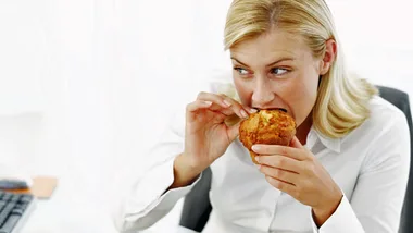Woman in white shirt eating a muffin at her desk while looking sideways.