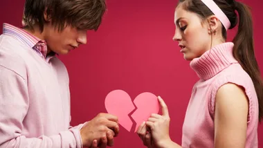 Young couple against a pink background holding broken heart pieces, expressing relationship troubles.