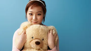 Young woman holding a large teddy bear against a blue background, smiling at the camera.