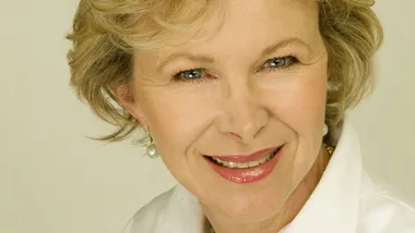 Portrait of a smiling woman with short, blonde hair and pearl earrings against a light background.