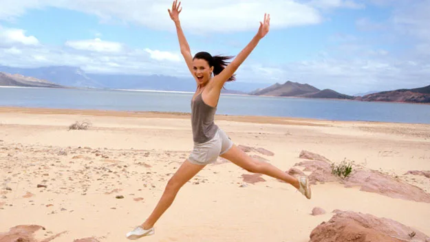 A woman joyfully jumping on a sandy beach, arms raised, with mountains and blue sea in the background.