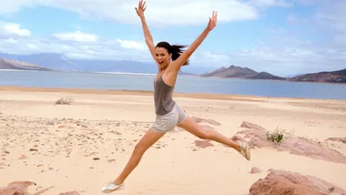 A woman joyfully jumping on a sandy beach, arms raised, with mountains and blue sea in the background.