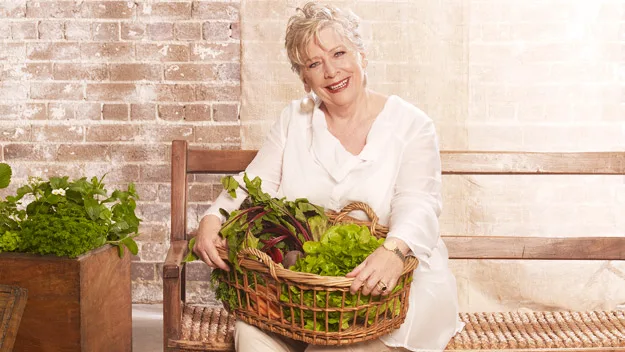 A woman in white holding a basket of greens, seated on a bench with a brick wall background.
