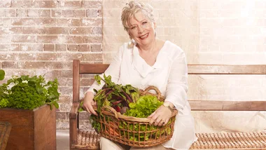 A woman in white holding a basket of greens, seated on a bench with a brick wall background.