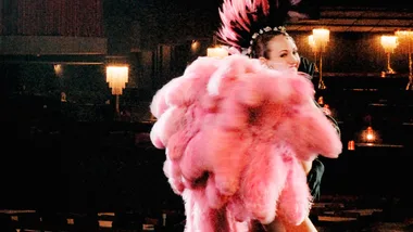 Showgirl performing with pink feathers on stage, dimly lit, wearing a headdress.