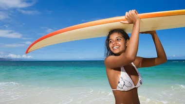 Young woman holding a surfboard on a sunny beach with clear skies and turquoise water.