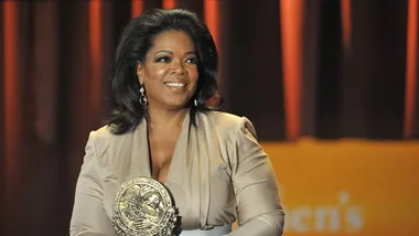 Oprah Winfrey smiling, holding a prestigious award, with a warmly lit stage backdrop.