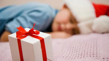 Child wearing Santa hat sleeping beside gift box with red ribbon.