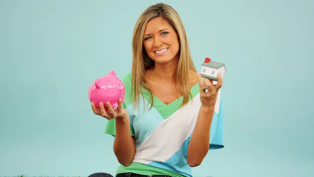 Person holding a pink piggy bank and a small model house, smiling against a light blue background.