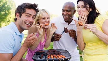 Group of four friends enjoying grilled food at a barbecue outdoors on a sunny day.