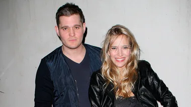 A couple stands close together, both wearing casual dark clothing, smiling against a plain backdrop.