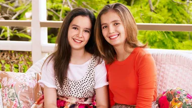Two smiling young girls sitting on a wicker couch outdoors, one in a polka dot dress and the other in an orange top.