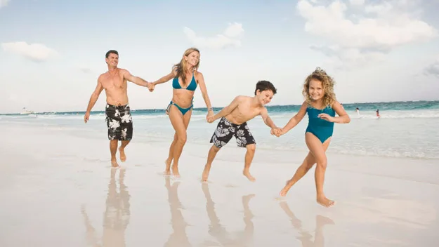 Family of four playing and holding hands on a sunny beach with blue sky and ocean in the background.