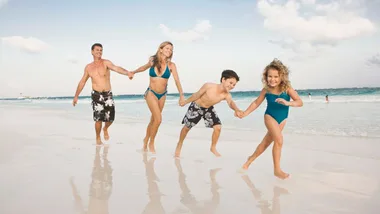 Family of four playing and holding hands on a sunny beach with blue sky and ocean in the background.
