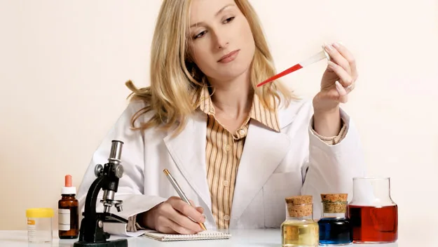 Scientist in a lab coat examining a red liquid in a pipette, with a microscope and glass bottles on the table.