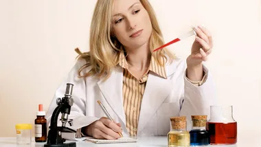 Scientist in a lab coat examining a red liquid in a pipette, with a microscope and glass bottles on the table.