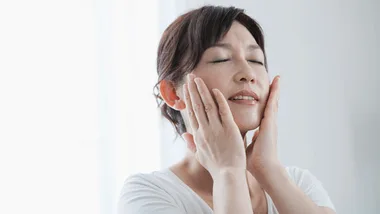 A woman performing facial exercises with eyes closed, gently massaging cheeks, in a bright room.
