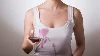 A person holding a glass of red wine with a spill on their white tank top.