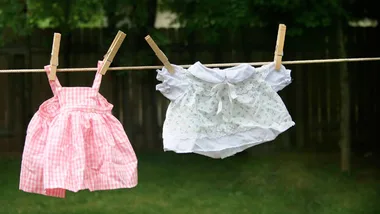 Children's clothes hang on a line: pink plaid dress and white floral top drying outdoors.