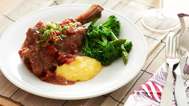 Braised lamb shank with polenta, topped with herbs, served alongside steamed broccoli on a white plate.