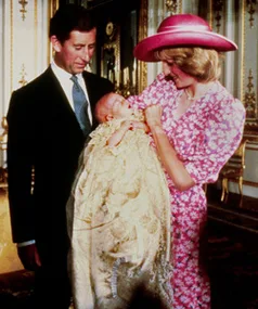 A couple with a baby in a christening gown; the woman wears a pink dress and matching hat, standing together in a formal setting.