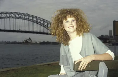 Young woman with curly hair sitting on a bench, Sydney Harbour Bridge and Opera House in the background.