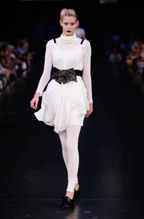 Model on runway in white dress with black belt, wearing black lipstick, and white leggings against a dark background.
