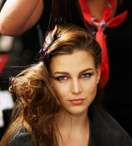 Model with styled hair being prepared, wearing a cape, focused expression.