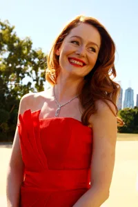 Red-haired woman in a strapless red dress smiles brightly outdoors on a sunny day.