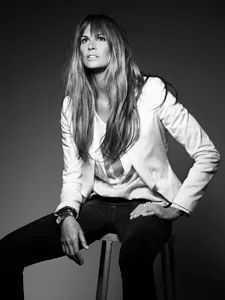 Black and white photo of a woman with long hair, seated, wearing a white blazer and dark pants, looking upward.