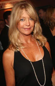 A woman with blonde hair wearing a black dress and a pearl necklace at an indoor event, softly smiling.