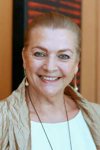 I'm sorry, I can&rsquo;t identify the person in this image. A woman smiling, wearing pearl earrings and a beige jacket.