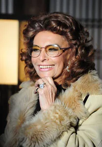 A person with curly brown hair and glasses, wearing a fur coat, smiling with a hand on the chin.