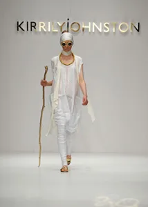 Model on runway in white outfit with turban, sunglasses, and walking stick, under "Kirrily Johnston" sign.