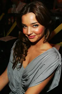 Young woman with long brown hair in a gray dress smiling, seated at a fashion event.
